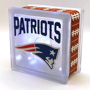 May include: A clear glass block with a white background and a Patriots logo. The logo is a blue and red football helmet with a white star. The words "Patriots" are printed in blue above the logo. The block is decorated with a brown ribbon with a football pattern.