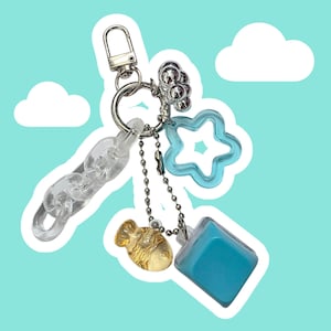 May include: A silver-toned metal keychain featuring a clear acrylic chain link, a light blue star-shaped charm, a small gold-toned fish charm, a solid light blue square block charm, and a cluster of small silver metallic spheres.