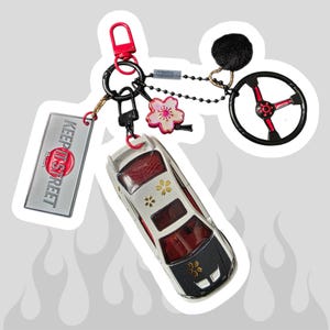 May include: A keychain featuring a white toy car with a carbon fiber hood, gold cherry blossom decals, and red interior. It includes a rectangular tag with the text KEEP IT STREET, a black steering wheel charm, a pink flower charm, and a black pom-pom.