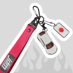 Peut inclure: Une sangle de porte-clés en tissu rouge avec un écusson rectangulaire noir portant le texte blanc JDM RACING. La sangle est fixée à un mousqueton en métal noir, une voiture miniature blanche et un petit pendentif rectangulaire au drapeau japonais.