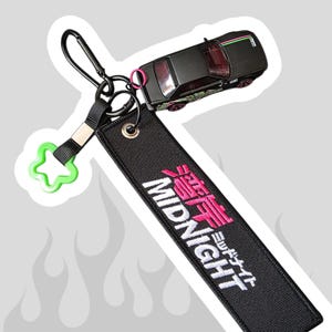 以下が含まれることがあります： MIDNIGHTと白とピンクの日本語で刺繍された黒い布製キーホルダー。黒い金属製カラビナ、小さなライムグリーンの星型プラスチックチャーム、ピンクのホイールアクセントが付いた黒いミニカーが取り付けられています。