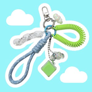 May include: A keychain featuring a blue braided cord, a lime green coiled wristlet, a clear plastic chain link, a small lime green square charm, and two iridescent cloud-shaped beads attached to a silver metal lobster clasp and ring.