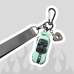 Puede incluir: Un llavero con una correa de tela gris oscuro sujeta a un mosquetón negro. Incluye un coche de juguete de color verde menta claro con una franja de carreras negra y el texto Mazda 5, junto con un pequeño dije metálico con forma de rotor.