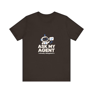 May include: A dark brown short-sleeve t-shirt featuring a graphic of a white and blue robot holding a speech bubble with a checkmark. Below the robot, the text reads ASK MY AGENT in large letters and I already delegated it. in smaller text.