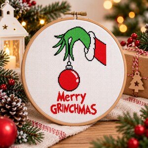 Puede incluir: Un bastidor de bordado circular de madera que contiene un diseño de punto de cruz sobre tela blanca. El diseño presenta una mano verde con una manga roja sosteniendo un adorno navideño rojo, con el texto rojo Merry GRINCHMAS debajo.