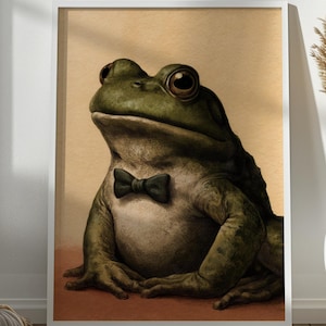 May include: A framed wall art print featuring a detailed illustration of a green frog wearing a small black bow tie. The frog is positioned against a solid tan background, with its large dark eyes looking forward in a seated, upright posture.
