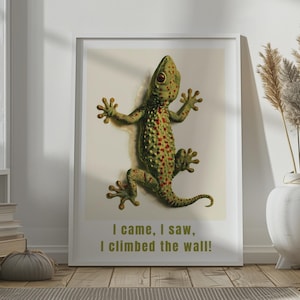 May include: A framed wall art print featuring a green gecko with red spots on a light background. The gecko is depicted in a climbing position. Below the gecko, the text reads: I came, I saw, I climbed the wall! The print is in a white frame.