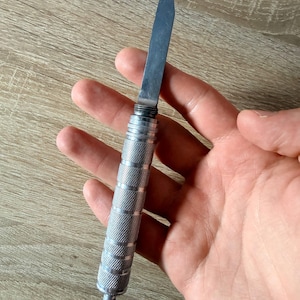 Può includere: Un coltello da precisione color argento con una lama in metallo sottile e appuntita e un manico cilindrico caratterizzato da una texture zigrinata a incrocio. L'utensile presenta una finitura metallica e una piccola sporgenza arrotondata alla base.