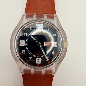 Op de afbeelding: Een Swatch-polshorloge met een doorzichtige kunststof behuizing en een oranje siliconen horlogeband met textuur. De zwarte wijzerplaat heeft witte Arabische cijfers op 12, 9 en 6, een dag-datumvenster op 3 uur met WED 1 en zilverkleurige wijzers.