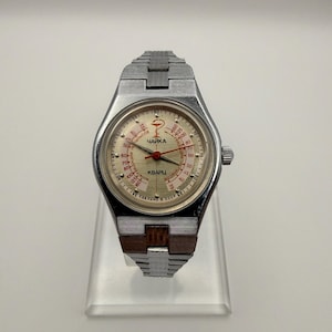 May include: A silver-tone wristwatch with a metallic link band featuring a light-colored dial. The face displays the text "ЧАЙКА" and "КВАРЦ" with a medical symbol, red hands, and a scale printed in red and black. The bottom edge reads "СДЕЛАНО В СССР".