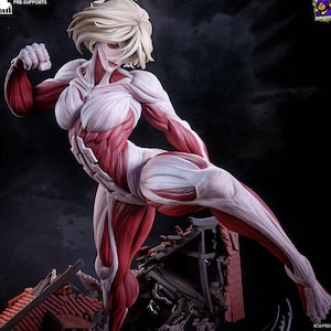 Pode incluir: Uma estatueta detalhada de uma titã feminina com cabelo loiro, representada com tecido muscular vermelho exposto e tecido conjuntivo branco. A figura está numa pose dinâmica de pontapé sobre um edifício em miniatura com telhado vermelho.