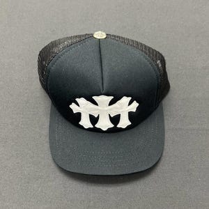 Könnte beinhalten: Eine schwarze Trucker-Cap mit einer einfarbig schwarzen Vorderseite und einer weißen, kreuzförmigen Applikation in der Mitte. Die Kappe verfügt über ein schwarzes Netzgewebe an der Rückseite, einen flachen Schirm und einen kleinen silbernen Knopf.