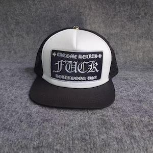 May include: A black and white trucker cap featuring a white foam front panel with a black rectangular patch. The patch displays the text Chrome Hearts, FUCK, and Hollywood, USA in a gothic font. The cap has a black brim and mesh back.
