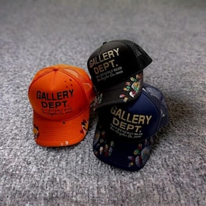 May include: Three trucker hats in orange, black, and navy blue. Each hat features the text GALLERY DEPT. 7613 BEVERLY BLVD Los Angeles Ca. 90036. The orange hat has white paint splatters, while the black and navy hats have colorful paint accents.