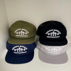 May include: Four baseball caps stacked in two columns, featuring olive green, black, navy blue, and light grey colours. Each cap displays white embroidered text reading CHROME HEARTS HOLLYWOOD with cross symbols and a decorative rope across the brim.