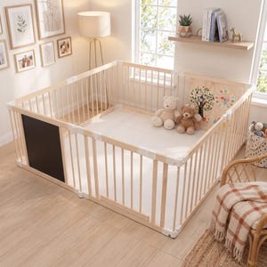 May include: A light-colored wooden playpen featuring vertical slats, white plastic corner connectors, and a black chalkboard panel on one side. Inside, a white floor mat holds two plush teddy bears and a wooden panel with a colorful tree graphic.