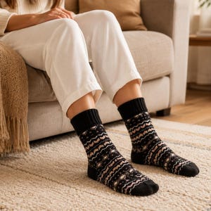 May include: A pair of mid-calf socks featuring a black base with a repeating geometric pattern in shades of cream and soft pink. The socks have a solid black ribbed cuff at the top and a solid black toe section, providing a contrast to the intricate knit design.