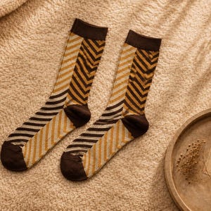 May include: A pair of crew socks featuring a split design with a dark brown cuff, heel, and toe. The leg and foot sections display a vertical split pattern, with one side showing a mustard yellow and white chevron print and the other side featuring dark brown and white diagonal stripes.