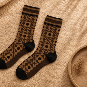 Puede incluir: Un par de calcetines de media caña con un patrón geométrico repetitivo de flores de color marrón oscuro sobre un fondo marrón claro. Los calcetines tienen talones y punteras de color negro sólido, con puños acanalados a rayas.