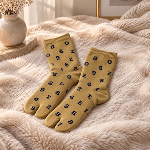 May include: A pair of mustard yellow tabi socks featuring a repeating pattern of dark navy blue numbers and symbols. These split-toe socks have a ribbed cuff and a distinct separation between the big toe and the other toes.