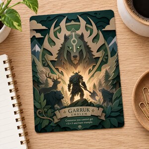 May include: A rectangular card featuring a layered paper-cut style illustration of a warrior holding an axe, surrounded by a wolf, a deer, and a bear. The text reads GARRUK EMBLEM and Creatures you control get +3/+3 and have trample.