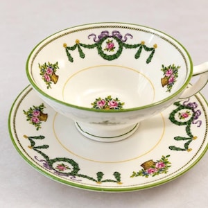 May include: A white porcelain teacup and saucer set featuring a green rim, a central floral bouquet of pink roses, and decorative swags of green garlands with purple ribbons and small yellow accents printed around the interior and exterior.