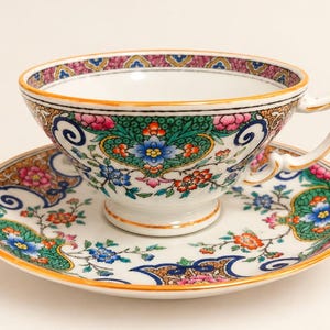 May include: A white porcelain teacup and saucer set featuring a vibrant, multi-colored floral pattern with green, blue, pink, and orange accents. The rim of the cup and saucer is decorated with a gold-trimmed border and intricate scrollwork.