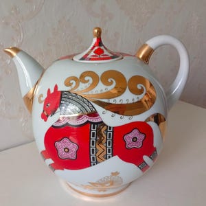 May include: A white porcelain teapot featuring a vibrant red horse illustration with gold scrollwork accents. The teapot has a matching lid with a red floral motif, a curved handle, and a spout with gold trim. The body displays geometric patterns.