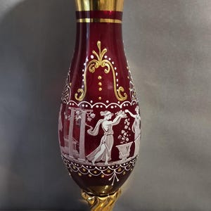 Puede incluir: Un jarrón de cristal rojo intenso con un borde dorado acampanado y una base de pedestal a juego con un tallo retorcido. El cuerpo del jarrón está decorado con figuras de esmalte blanco con vestimenta clásica, columnas arquitectónicas y filigranas doradas.