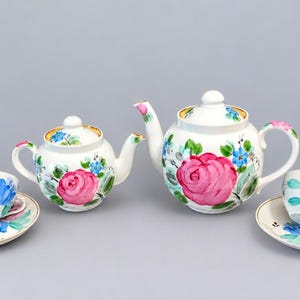 Pode incluir: Um conjunto de chá em cerâmica branca composto por dois bules e duas chávenas com pires a condizer. Cada peça é decorada com rosas cor-de-rosa, flores azuis e folhas verdes pintadas à mão. Os bules têm tampas com bordos dourados.