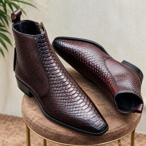 Puede incluir: Un par de botines con un patrón de textura de piel de serpiente en color marrón oscuro. Los botines tienen punta afilada, cierre de cremallera lateral con tirador dorado, una trabilla en el talón y suela negra con tacón bajo.