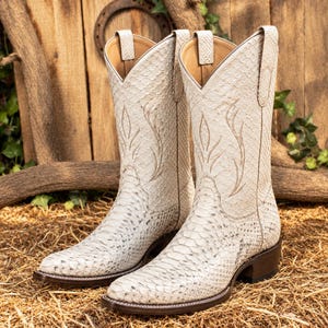 May include: A pair of off-white cowboy boots featuring a textured snakeskin pattern, pointed toes, and decorative stitching on the shaft. The boots have a stacked wooden heel, a dark brown sole edge, and pull tabs at the top of the boot opening.