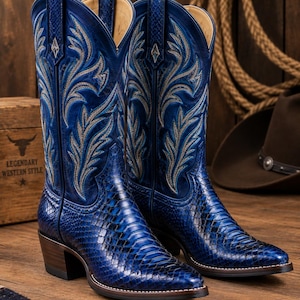 Puede incluir: Un par de botas vaqueras azul profundo con un pie de cuero texturizado con patrón de piel de serpiente y una caña alta con intrincados bordados plateados. Las botas tienen punta afilada, tacón de madera y un emblema metálico en la parte superior.