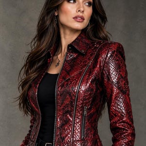 Peut inclure: Une veste ajustée présentant un motif peau de serpent rouge foncé et noir, un col pointu, des manches longues et une fermeture éclair frontale. Le vêtement comprend deux poches zippées verticales sur le devant et une ceinture à boucle métallique argentée.