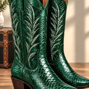 May include: A pair of emerald green cowboy boots featuring a textured snakeskin-like pattern and white stitched leaf-shaped embroidery on the shaft. The boots have a pointed toe, a stacked wooden heel, and pull tabs at the top of the shaft.