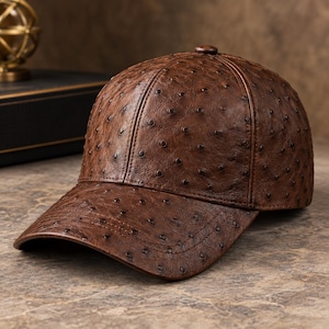 Puede incluir: Una gorra de béisbol marrón con un patrón texturizado inspirado en el avestruz, que presenta pequeñas hendiduras circulares oscuras en toda la superficie. La gorra tiene una corona estructurada, una visera curva y un botón central a juego.