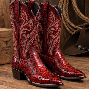 May include: A pair of red cowboy boots featuring a textured snakeskin pattern, a pointed toe, and a stacked heel. The upper shaft of each boot is decorated with intricate cream-colored stitching in a leaf-like design and includes pull tabs.