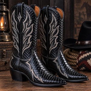 Op de afbeelding: Een paar zwarte cowboylaarzen met een gestructureerd slangenleerpatroon en ingewikkeld zilverkleurig borduurwerk in een vlamachtig ontwerp op de schacht. De laarzen hebben een puntige neus, een blokhak en treklipjes aan de bovenkant.