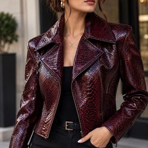 Può includere: Una giacca da motociclista color bordeaux intenso con una texture lucida in pelle di serpente, un ampio colletto dentellato e una chiusura con cerniera in metallo argentato. La giacca è indossata sopra un top nero e pantaloni neri con cintura.