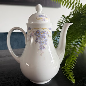 Op de afbeelding: Een witte porseleinen theepot met een delicaat bloemmotief van kleine blauwe bloemetjes, groene blaadjes en dunne gele decoratieve lijnen. De theepot heeft een gebogen handvat, een lange tuit en een bijpassend deksel met een ronde knop.