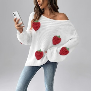 Puede incluir: Un suéter de punto blanco de manga larga con escote ancho y un patrón de fresas texturizado. El suéter presenta aplicaciones tridimensionales de fresas rojas con hojas verdes distribuidas por el frente y las mangas, combinado con pantalones vaqueros azules.