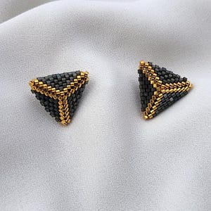 Op de afbeelding: Een paar driehoekige oorknopjes gemaakt van kleine, vierkante kraaltjes. De oorbellen hebben een donkergrijze kleur met metallic gouden kraalaccenten die een duidelijk Y-vormig patroon vormen langs de randen en het midden.