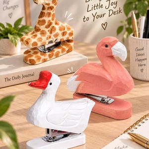 May include: Three animal-shaped staplers featuring a carved wooden texture. One is a brown and tan giraffe, one is a pink flamingo with a white beak, and one is a white goose with a red beak. Each stapler has a metal mechanism at the base.