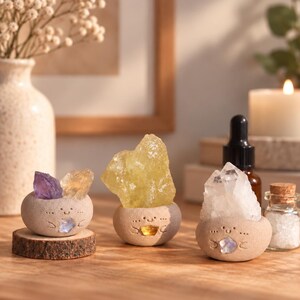 May include: Three small, round, beige ceramic planters with carved smiling faces, each holding raw crystals. One holds purple and yellow stones, another a large yellow crystal, and the third a clear quartz cluster with a small iridescent stone.