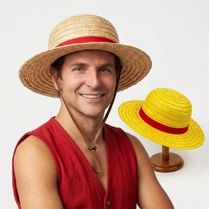 May include: A natural-coloured straw hat with a red ribbon band and a brown chin strap, alongside a bright yellow straw hat with a red ribbon band displayed on a wooden stand. Both hats feature a wide brim and a rounded crown design.
