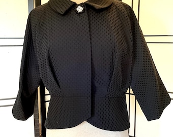 May include: A black, textured jacket with a round collar and three-quarter sleeves. The jacket features a decorative button and a fitted waist. The fabric has a subtle, repeating pattern.