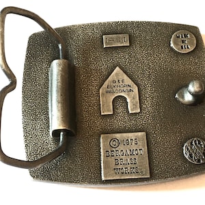 May include: A vintage metal belt buckle with a textured surface. The buckle features the text "I-51", "D&E ELKHORN WISCONSIN", "&copy; 1975 BERGAMOT BRASS WORKS", and "MADE IN USA". The buckle has a rectangular shape with a curved metal loop.