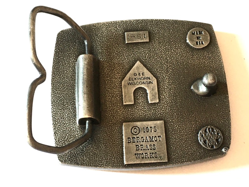May include: A vintage metal belt buckle with a textured surface. The buckle features the text "I-51", "D&E ELKHORN WISCONSIN", "&copy; 1975 BERGAMOT BRASS WORKS", and "MADE IN USA". The buckle has a rectangular shape with a curved metal loop.