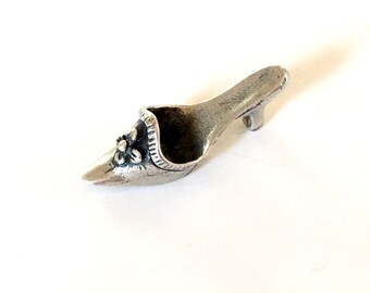 May include: A small, silver-coloured shoe figurine with a pointed toe and a small heel. The shoe features a decorative flower on the side and a detailed trim around the opening. The figurine is likely made of metal and is a decorative item.