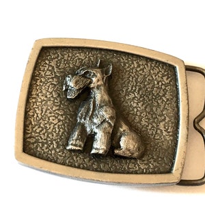 May include: A rectangular metal belt buckle featuring a textured background and a sculpted dog figure. The dog is sitting, and the buckle has a silver frame and a bronze clasp.